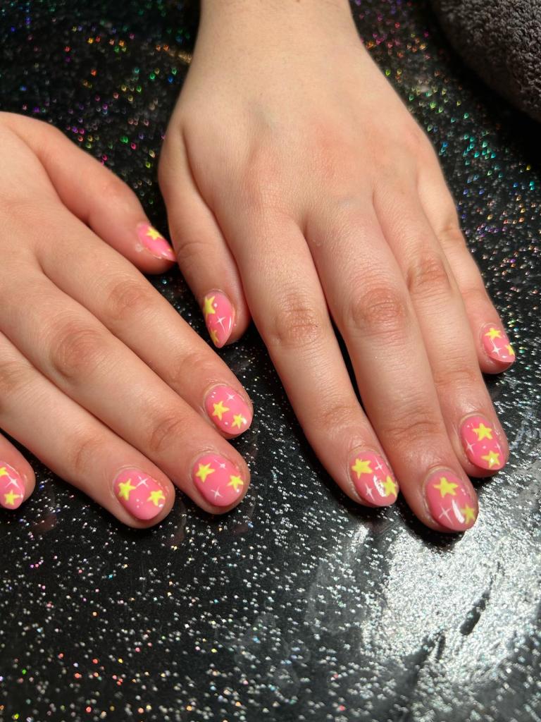 Close-up of hands with pink nail polish featuring yellow star designs, resting on a sparkling black surface.