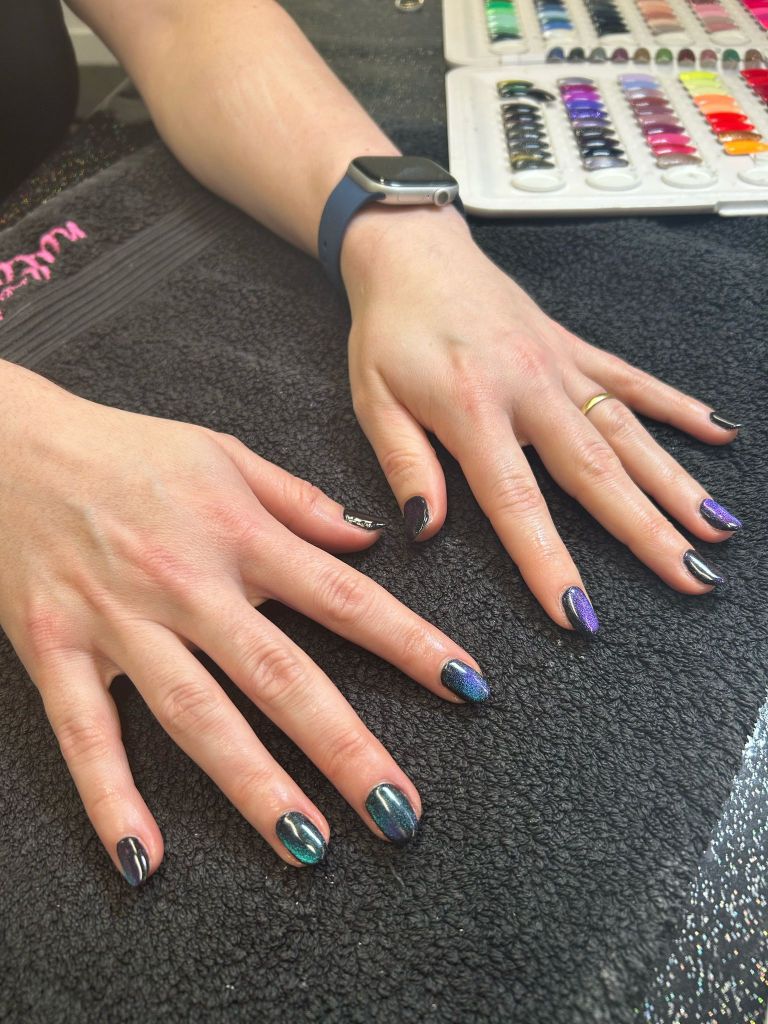 Close-up of hands displaying manicured nails in various shades of dark, shimmering colors, resting on a black surface with nail polish swatches in the background.