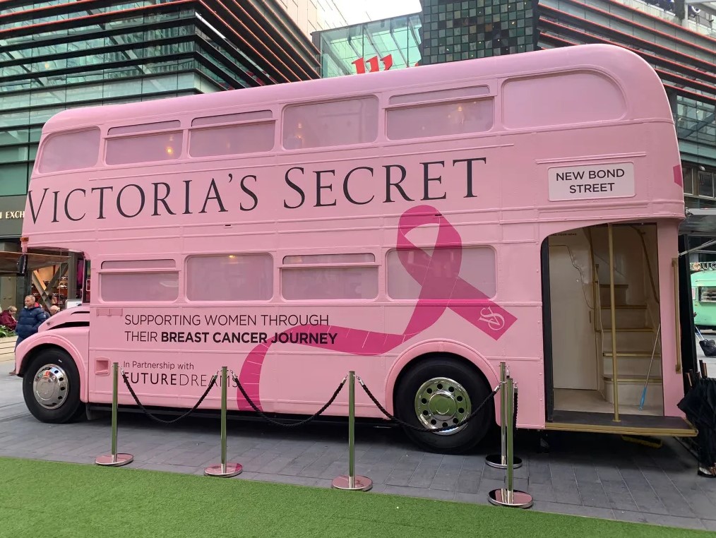 A pink double-decker bus branded with 'Victoria's Secret', featuring a pink ribbon symbol, parked in an urban area, promoting support for women's breast cancer journeys.