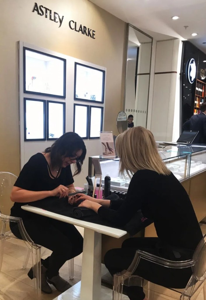 Two women sitting at a table in a nail salon, one is receiving a manicure while the other is working on her nails. The salon has a sign that reads 'ASTLEY CLARKE' in the background.