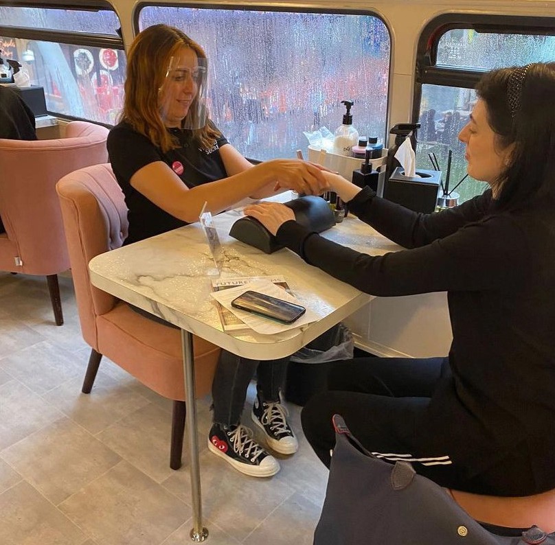 A nail technician in a black shirt and a face shield performs a manicure on a seated client inside a cozy setting, with a marble table and a rain-soaked window in the background.