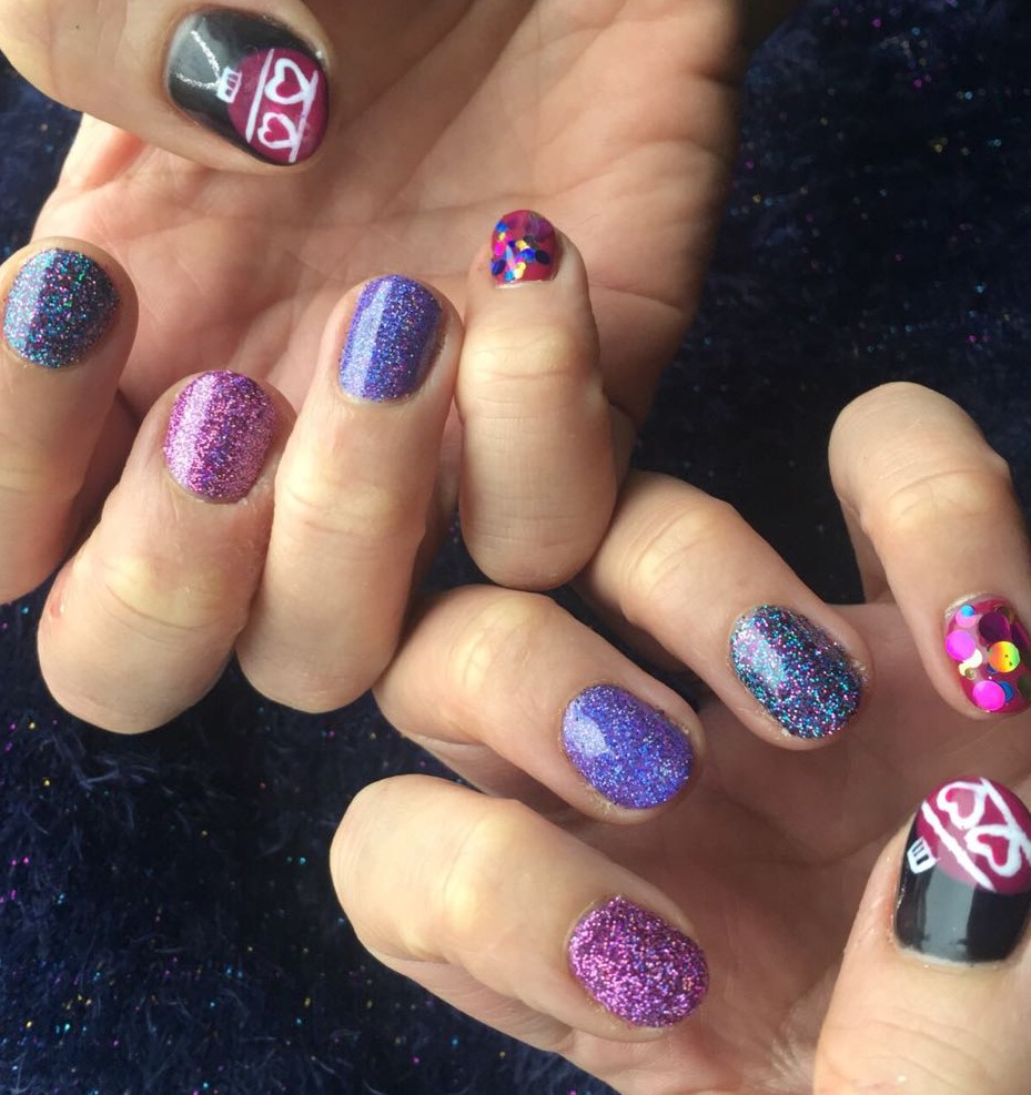 Close-up view of festive nail art featuring a variety of glittery and colorful designs, including purple and pink sparkles, and a whimsical graphic on one nail.