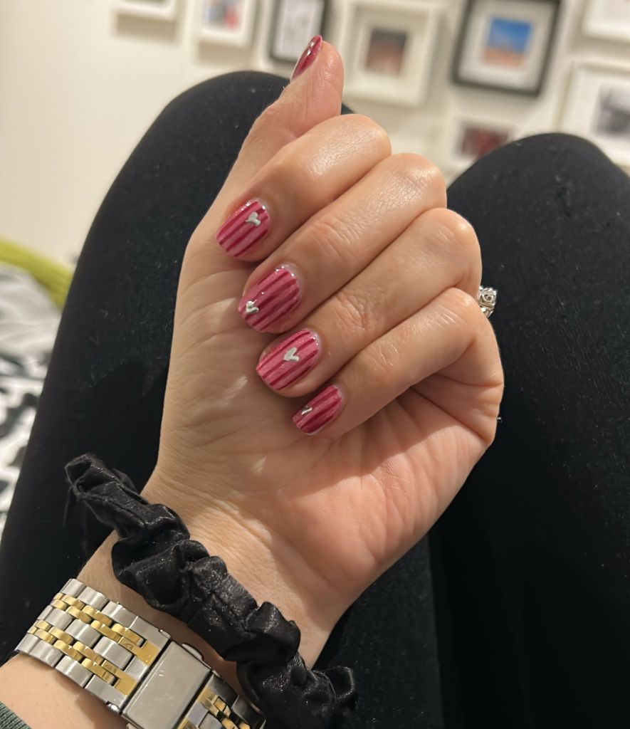 A close-up of a hand with manicured nails featuring a pink striped design with small white hearts. The wrist is adorned with a black scrunchie and a gold and silver watch.