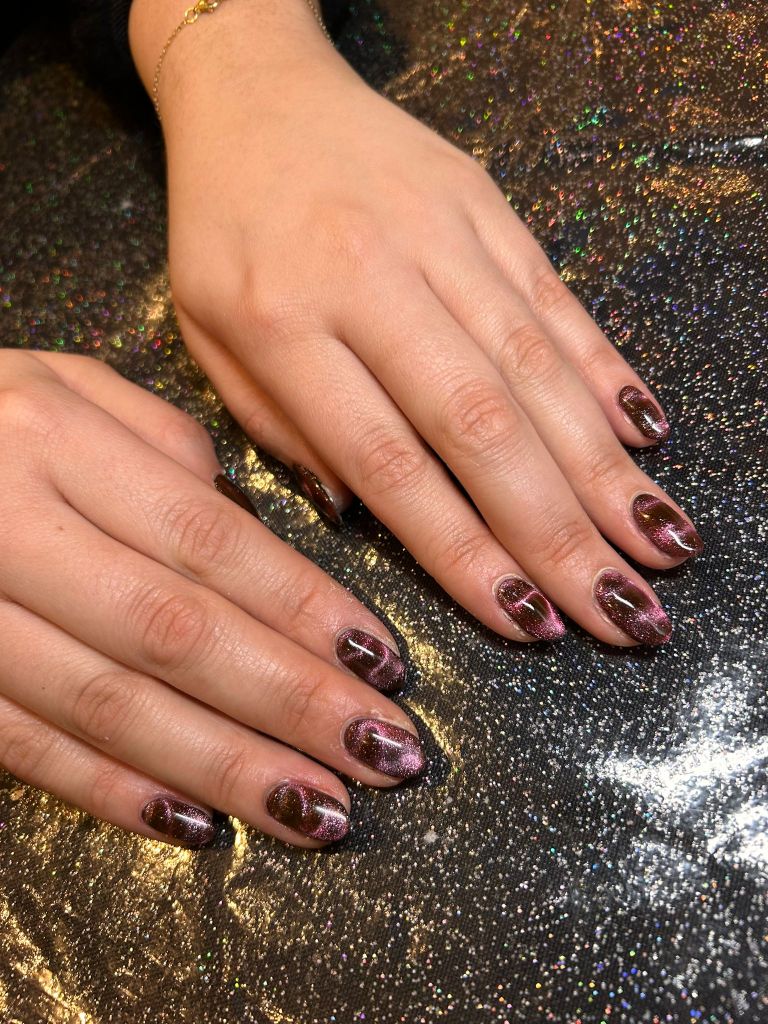 Close-up of two hands with stylish manicure featuring dark glittery nails with pink and purple ombre effects, resting on a sparkly background.