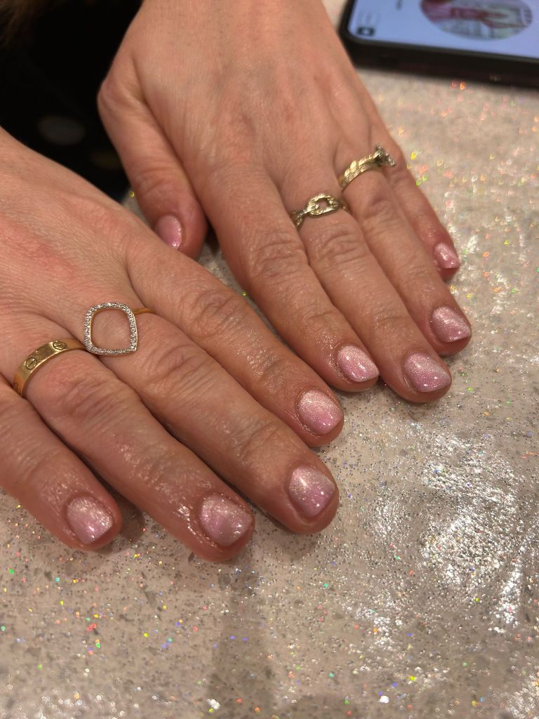 Close-up of two hands showing manicured nails painted with shimmering pink polish, adorned with several rings, resting on a sparkling surface.