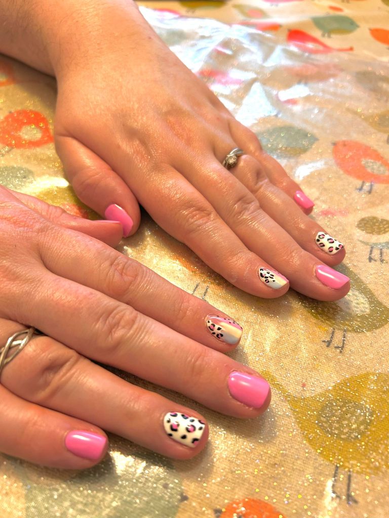 Close-up of two hands with manicured nails on a patterned tablecloth. The nails are painted in pastel pink with some accented nails featuring a white base with black spots and colorful designs.