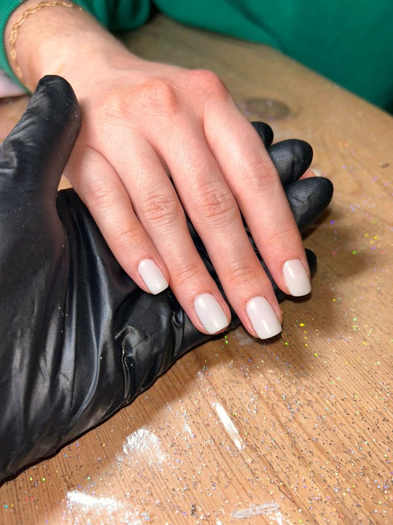 A close-up of a hand with neatly manicured light-colored nails resting on a wooden surface, while a gloved hand supports it.