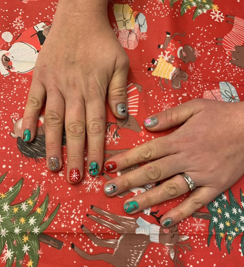 Christmas is coming! Two hands displaying festive Christmas-themed nail art on a red holiday tablecloth
